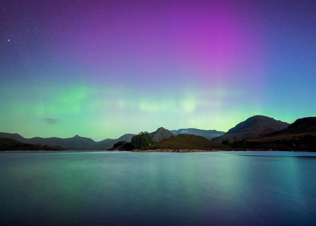 Purple and green aurora australis over a still lake in south-west Tasmania, with mountains and a forested island reflected in the calm water.
