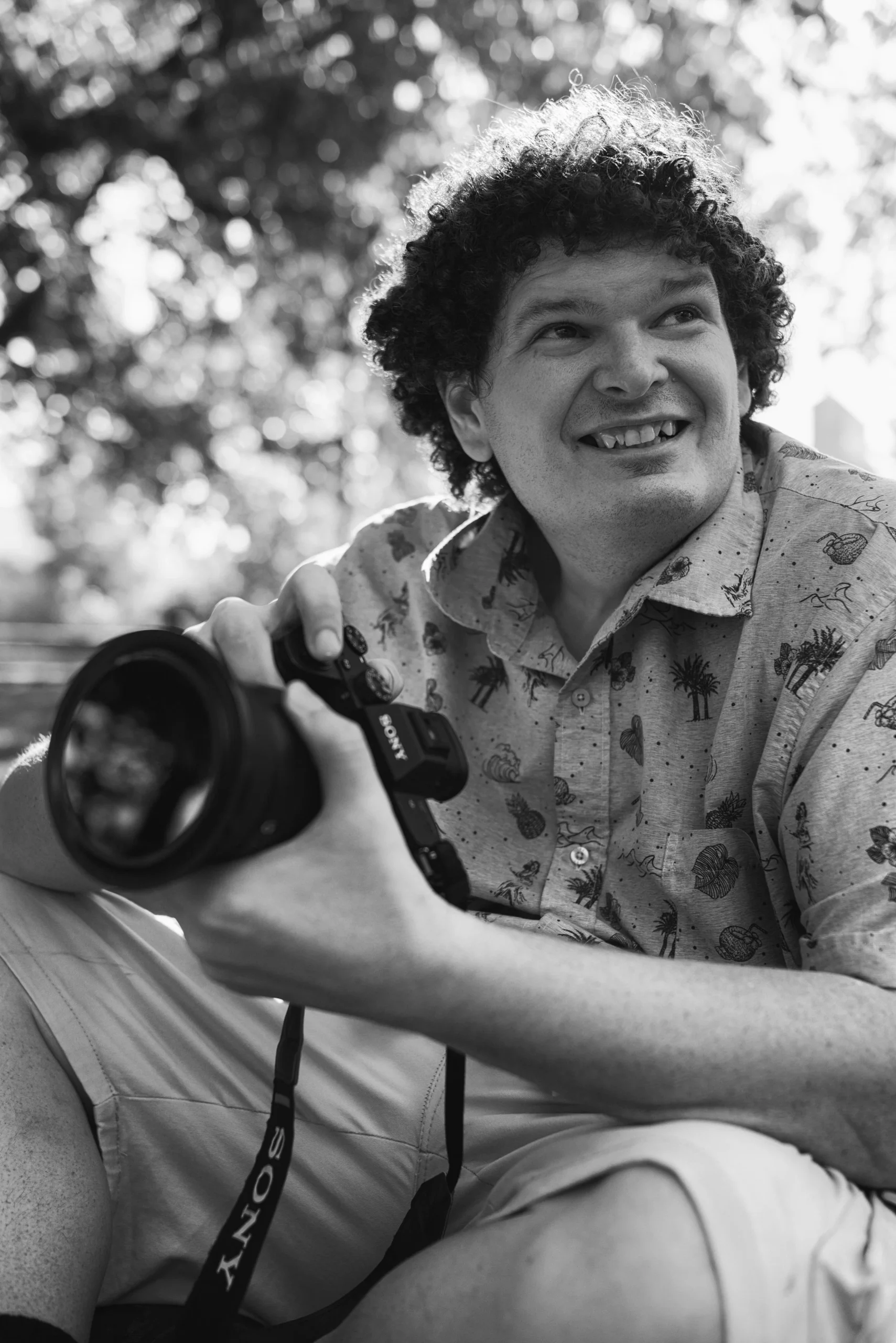 Ben is pictured with afternoon sun backlighting him and the trees above, while holding a camera and staring into the distance