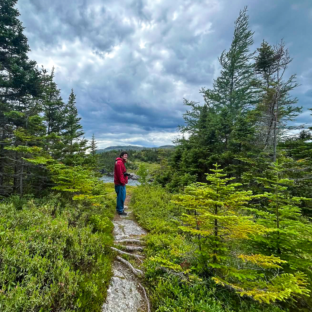 Ben is stood between encroaching coniferous trees beside a small lake, with mountains and foreboding skies behind.