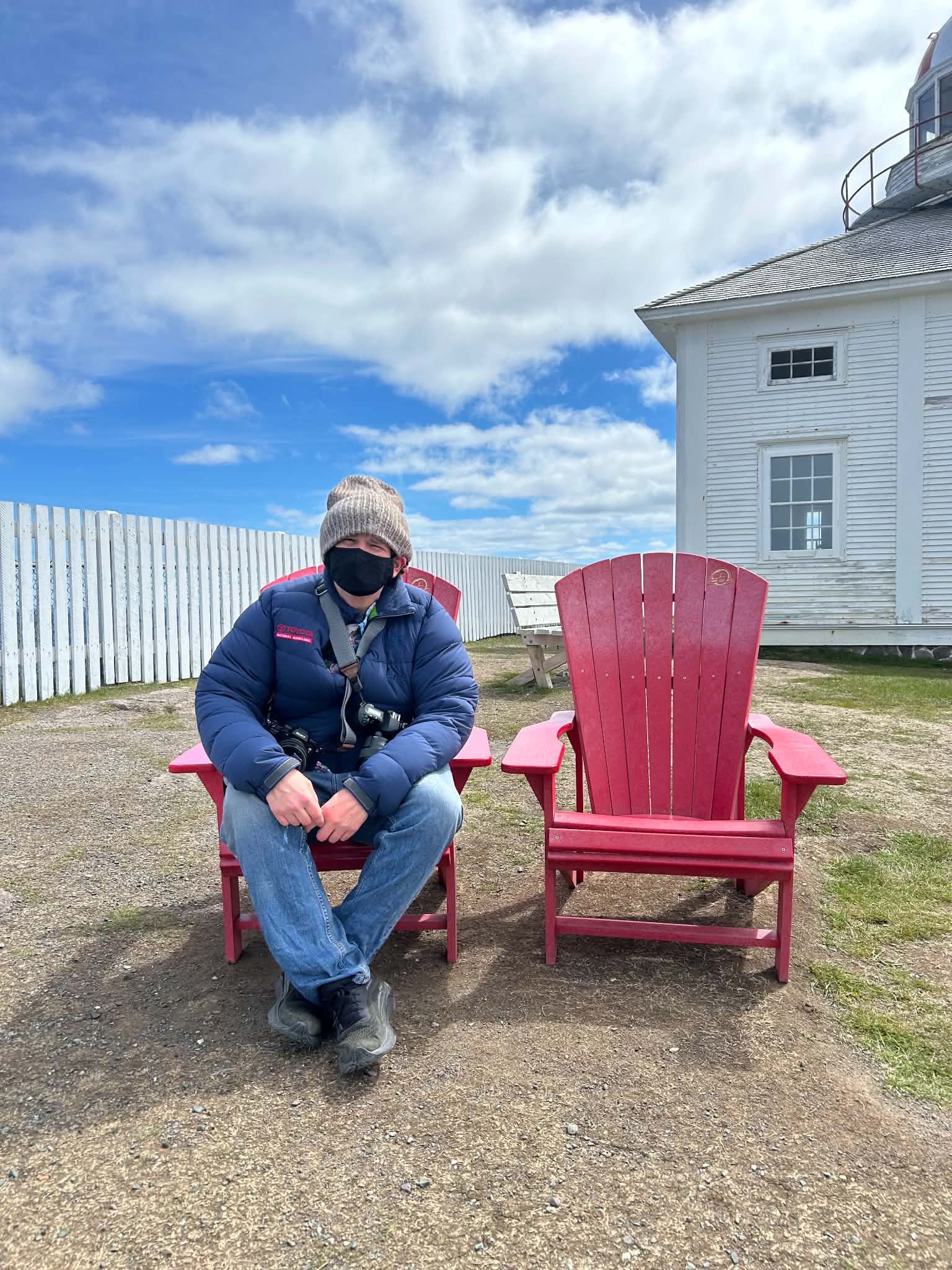 Ben is pictured, wearing full cold weather gear and a face mask while sitting in one of two red chairs beside Cape Spear Lighthouse